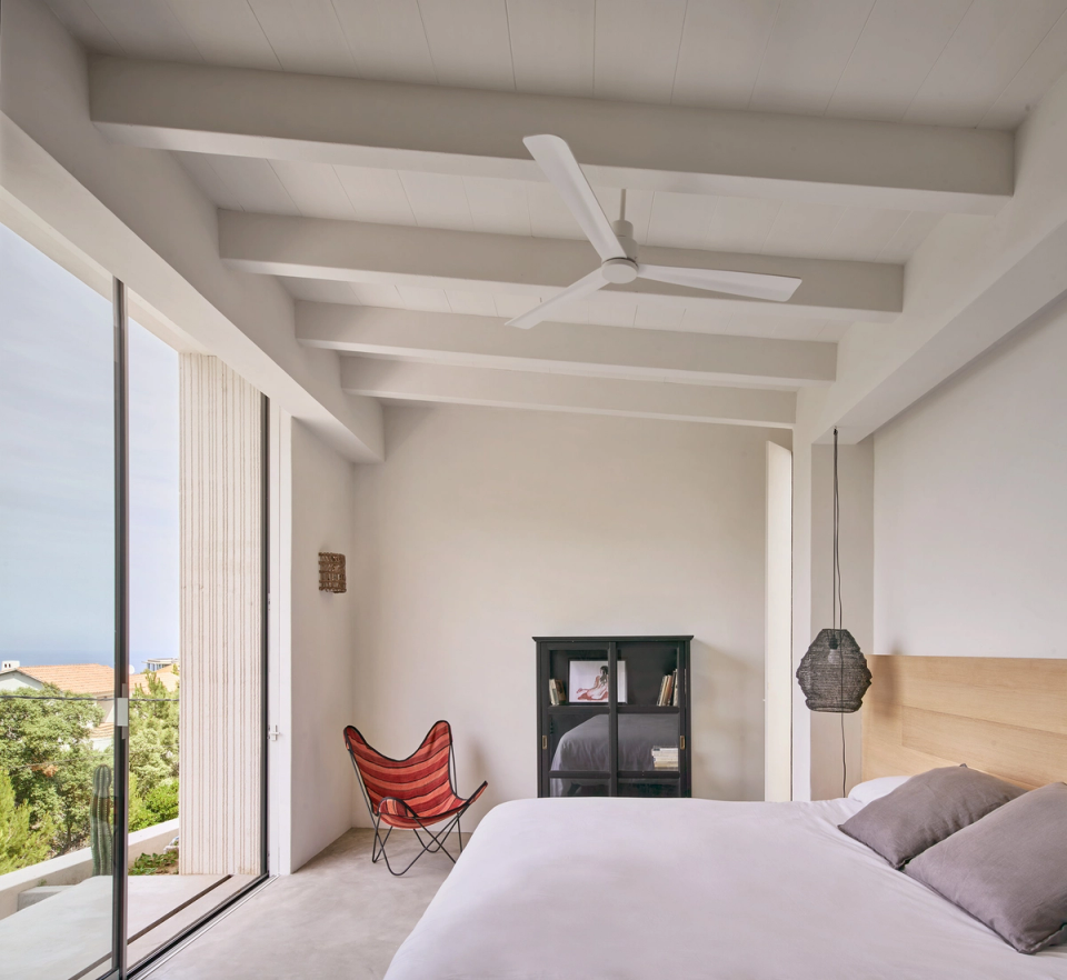 The master bedroom of this single-family home stands out for its minimalist architecture and direct relationship with the exterior through large panoramic openings. Natural light constantly enters the space, enhancing the feeling of calm and spaciousness, while neutral-toned finishes and natural wood reinforce a warm Mediterranean atmosphere. Exposed white beams and integrated furniture provide visual continuity and a clean, timeless architectural language. This single-family home prioritizes comfort, connection with the landscape, and an open, serene residential experience.