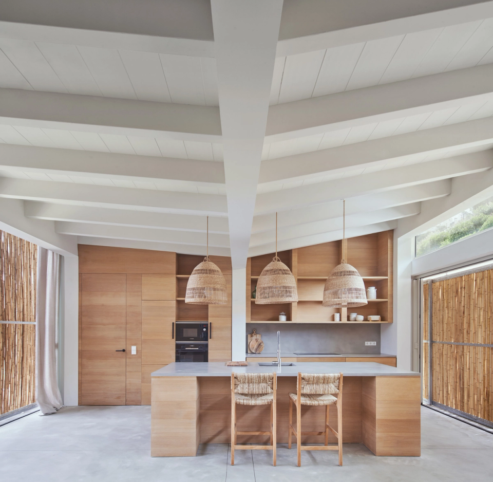 The kitchen of this single-family home integrates naturally into the main living space, embracing open, bright, and functional architecture. Large side openings allow a constant connection with the outdoors and enhance the entry of natural light throughout the day. Natural wood finishes, plant fibers, and the exposed roof structure reinforce the warm Mediterranean character of the home, creating a relaxed and elegant atmosphere. This single-family home stands out for its combination of noble materials, contemporary design, and spacious areas designed to encourage daily living and connection with the landscape.