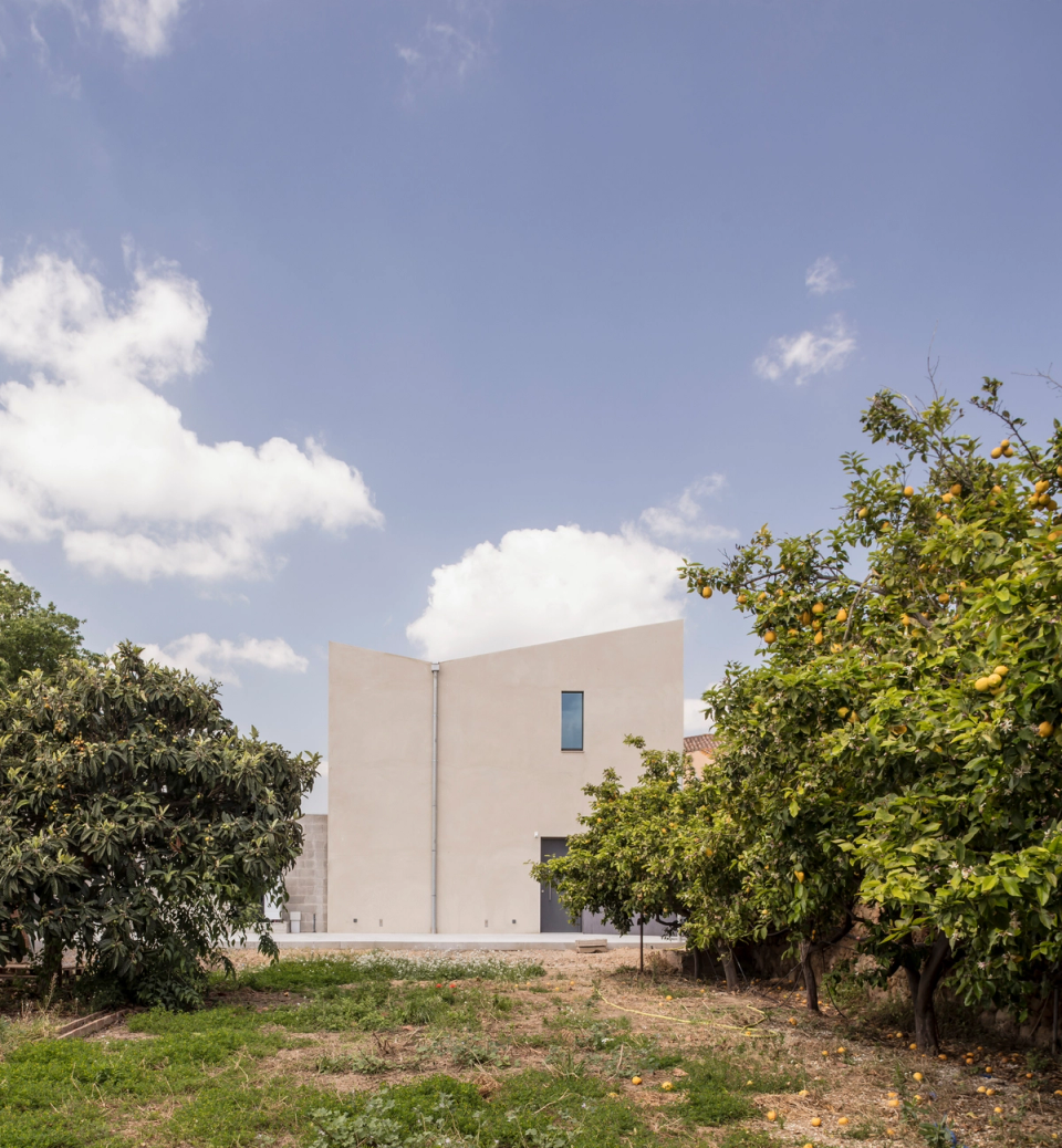 The rear façade integrates naturally into the surrounding vegetation, displaying a clean and abstract volumetry that dialogues with the landscape. Smooth surfaces and selective openings reinforce the formal simplicity of the house, while the presence of trees and natural light accentuate the relationship between architecture and nature.