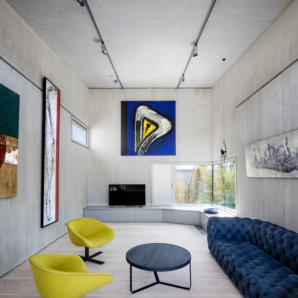 Interior of a contemporary single-family home with open spaces, generous ceiling heights, and a strong minimalist aesthetic. Exposed concrete finishes and large glazed openings provide spaciousness, natural light, and a direct connection with the outdoors. Designer furniture and contemporary art pieces introduce chromatic contrast and personality, creating a sophisticated, modern, and welcoming atmosphere within this architect-designed residential space.