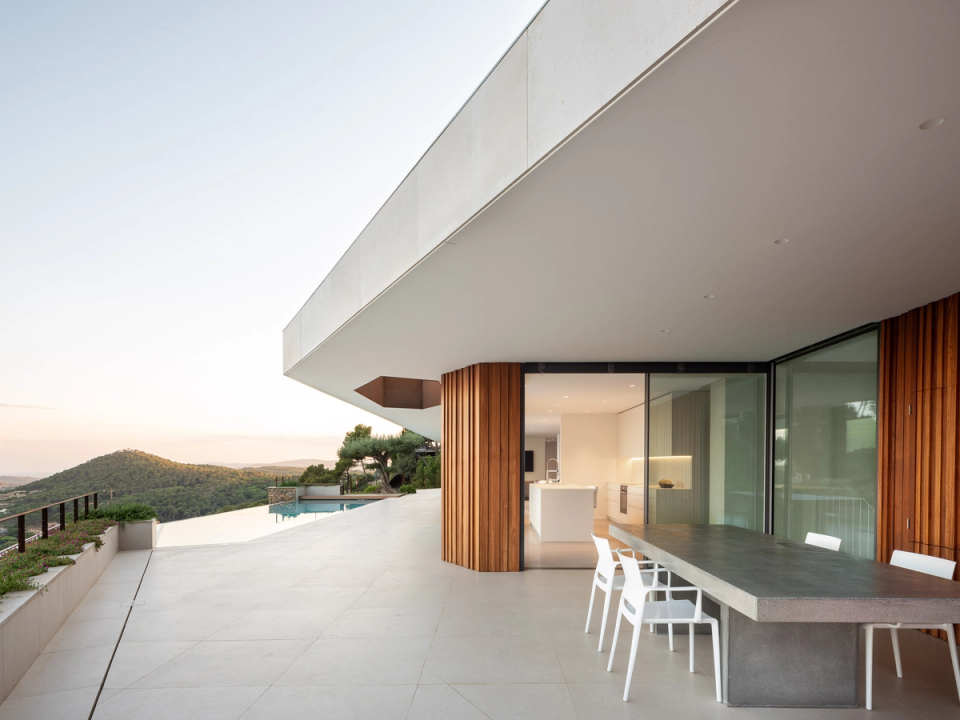 The detached single-family home unfolds along the slope with a highly refined contemporary architecture based on large horizontal planes of light concrete, natural stone walls, and vertical wood cladding that adds warmth. This image particularly highlights the large panoramic terrace facing the landscape, conceived as a direct extension of the interior spaces.
