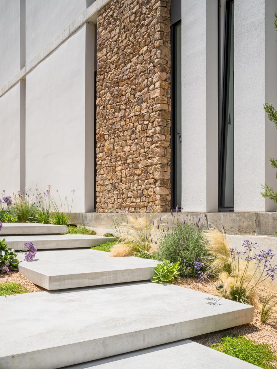 Detached single-family home with a façade combining natural stone, concrete, and minimalist finishes. Outdoor spaces and Mediterranean landscaping provide texture, natural character, and harmonious integration with the surroundings.
