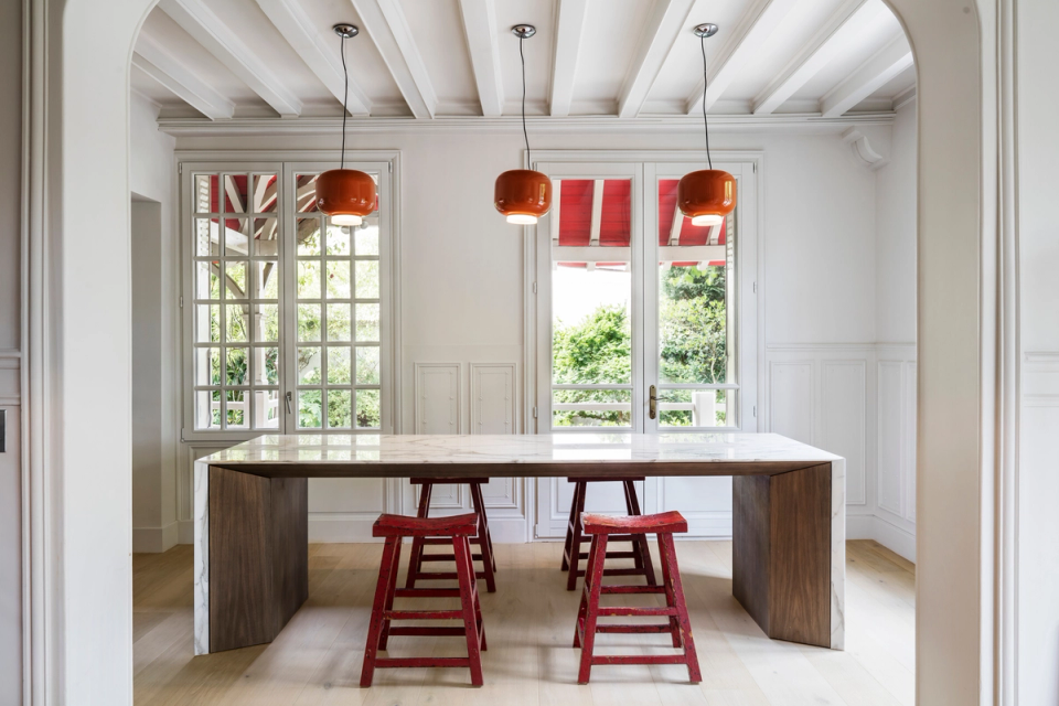 Interior renovation of an open kitchen that combines the elegance of classical architecture with contemporary statement pieces. Original moldings, exposed beam ceilings, and natural light interact with a large marble and wood island, while red-toned details add personality and contrast to a sophisticated and functional space.
