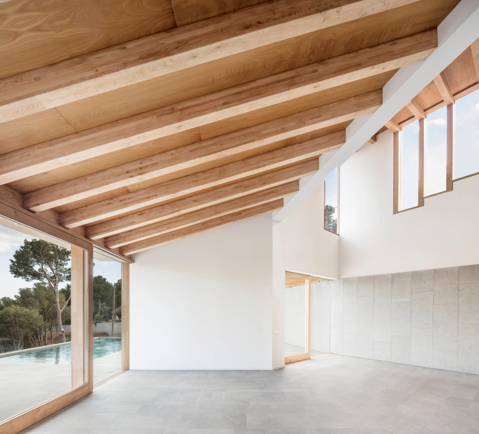 Interior of a detached single-family home with open spaces and architecture defined by sloped ceilings with large exposed wooden beams that provide warmth and character to the house. Large floor-to-ceiling openings create a direct connection with the outdoors and the swimming pool, enhancing natural light and the sense of continuity between interior spaces and the landscape.