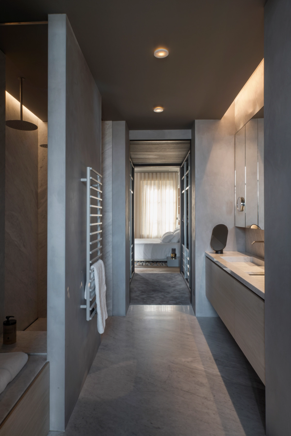 The interior renovation of this master bathroom embraces a refined and contemporary architecture, where continuous surfaces and a neutral color palette create an elegant and relaxing atmosphere. The linear layout visually connects the bathroom with the walk-in closet and bedroom, enhancing the feeling of spaciousness and continuity between spaces. Integrated lighting, suspended furniture, and minimalist finishes reinforce the sophistication of the design, transforming this space into an intimate and functional wellness area.
