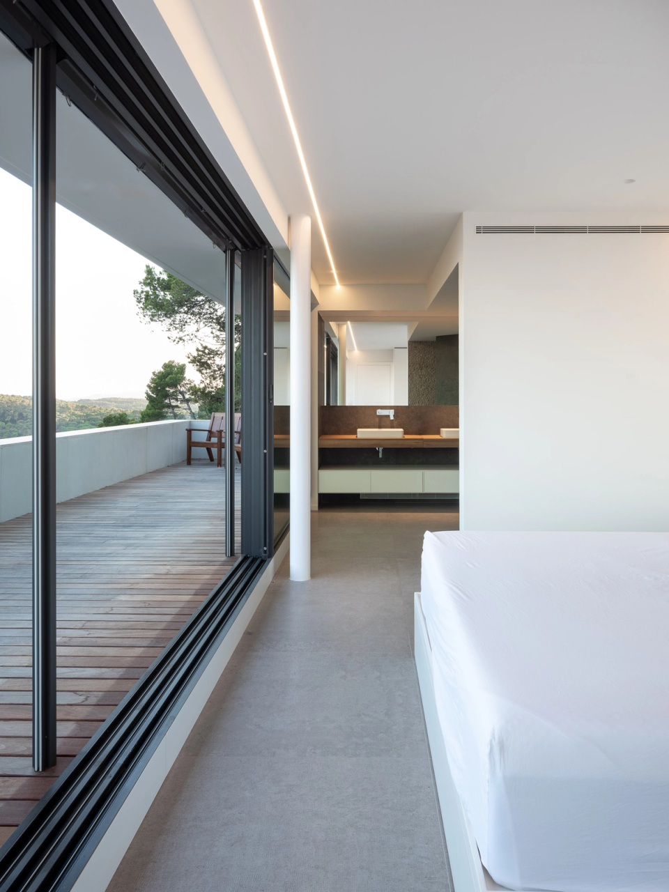 Master suite of a contemporary detached single-family home designed with an open architecture connected to the outdoors. Large sliding openings integrate the terrace and landscape into the interior space, enhancing natural light, views, and the sense of spaciousness. The combination of warm materials, minimalist lines, and integrated lighting creates an elegant, relaxing, and sophisticated atmosphere designed to prioritize comfort, functionality, and the constant relationship between interior and exterior.