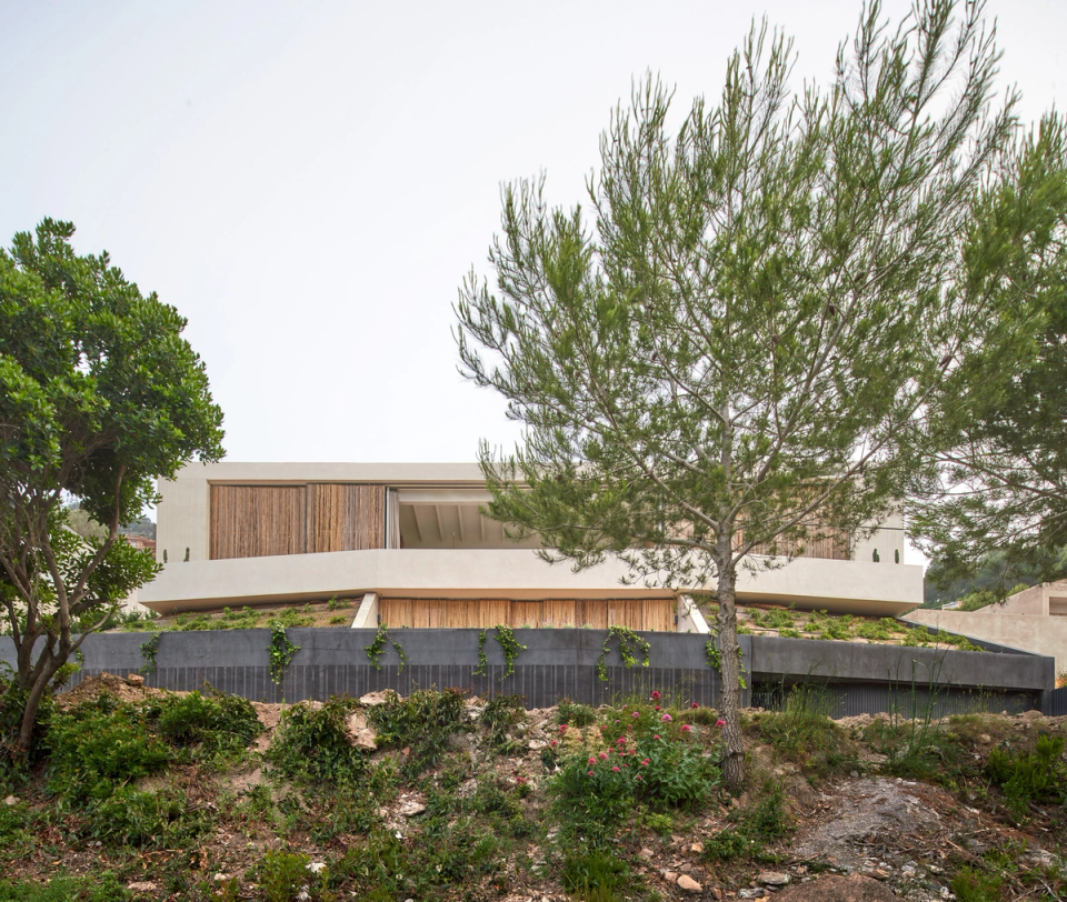 This single-family home blends sensitively into the natural landscape through architecture with soft geometries and organic materials. The project takes advantage of the site's topography to adapt to the surroundings and minimize visual impact, creating a balanced relationship between construction and nature. The light-colored façades, combined with wood cladding, provide warmth and a contemporary image that dialogues with the Mediterranean vegetation. This single-family home stands out for its discreet placement, green roofs, and an architectural concept designed to enhance light, privacy, and connection with the outdoors.
