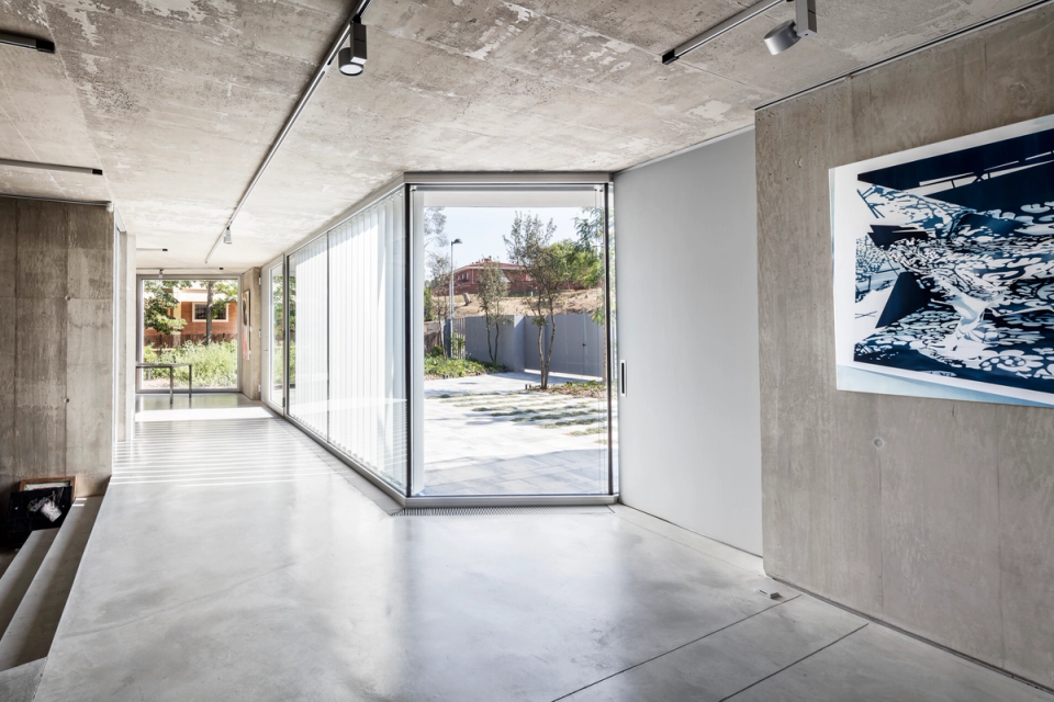 Interior of a contemporary single-family home with minimalist architecture based on the purity of materials and spatial continuity. Exposed concrete walls, continuous flooring, and large glazed enclosures define an elegant and functional space flooded with natural light. The direct relationship with the outdoors, combined with artistic elements and integrated technical lighting, brings character and sophistication to a modern and timeless architectural proposal.