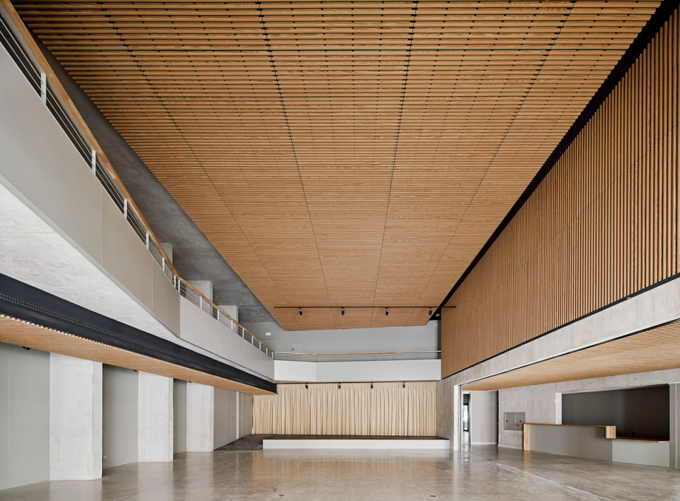 Architectural detail of the interior of a contemporary cultural center, where the combination of exposed concrete, metal structures, and wooden slat ceilings defines an elegant and functional space. The upper walkways and the great interior height provide visual dynamism and flexibility of use, allowing the cultural center to adapt to stage activities, exhibitions, and public events. The use of warm and industrial materials reinforces the modern identity of the building and creates a welcoming atmosphere designed for cultural and community life.