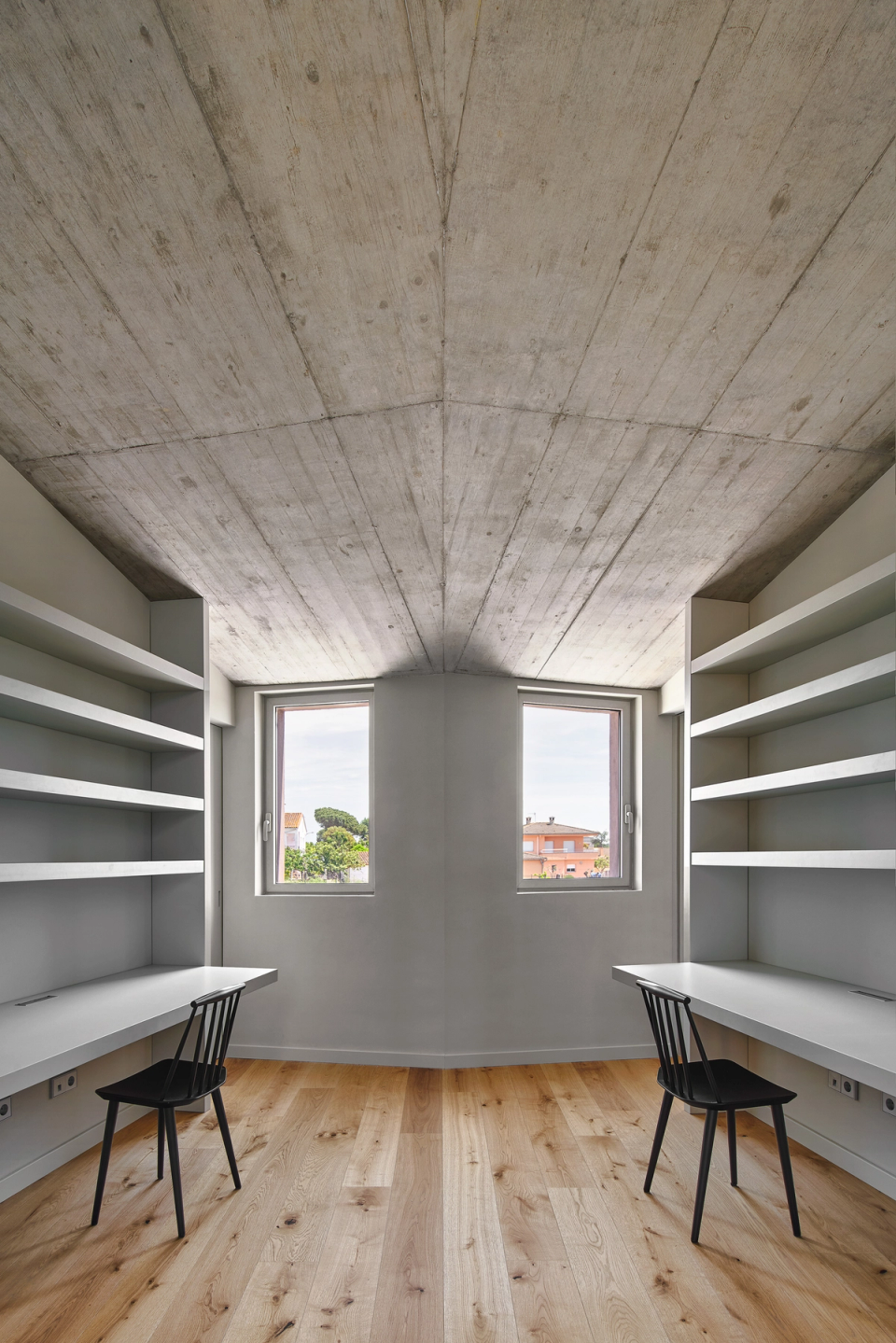 This workspace within the single-family home stands out for its minimalist and functional architecture, with integrated furniture and a symmetrical layout that enhances order and natural light. The exposed concrete ceiling and wooden flooring add personality and warmth to the interior.