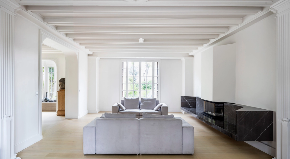 Interior renovation of a spacious and bright living room that merges classical architecture with contemporary design. Moldings, painted beam ceilings, and large openings are combined with minimalist furniture and a black stone-clad fireplace, creating an elegant, serene, and timeless space.