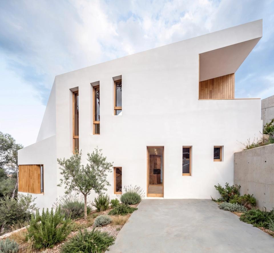 Detached single-family home with pure architectural lines and sculptural volumetry integrated into a Mediterranean natural setting. The white façade combines strategic vertical openings and natural wood cladding that provide warmth and visual contrast, reinforcing the project’s contemporary character. This detached house stands out for its natural light, connection with the landscape, and layout designed to adapt to the site’s topography. Landscaped outdoor spaces with native vegetation and noble materials contribute to creating elegant, sustainable architecture fully integrated with its surroundings.
