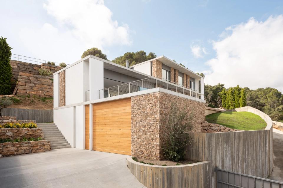 Detached single-family home distinguished by contemporary architecture combining geometric volumes, strong horizontal lines, and natural materials sensitively integrated into the environment. The natural stone façade and wooden cladding add warmth and texture, contrasting with the purity of white finishes and exposed concrete. The project adapts to the sloping site through retaining walls and outdoor platforms that connect the architecture with the surrounding Mediterranean landscape.