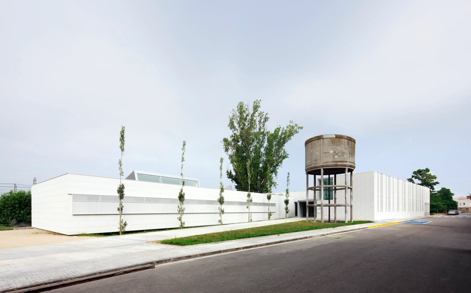 Exterior view of the Sant Vicenç Health Center, a contemporary healthcare facility with horizontal lines and minimalist architecture sensitively integrated into the urban surroundings. The complex stands out for its white façades, the brightness of its volumes, and the singular presence of the restored water tower, transformed into an iconic element of the project. The open and orderly outdoor space reinforces the sense of accessibility, calm, and functionality characteristic of a modern healthcare center.