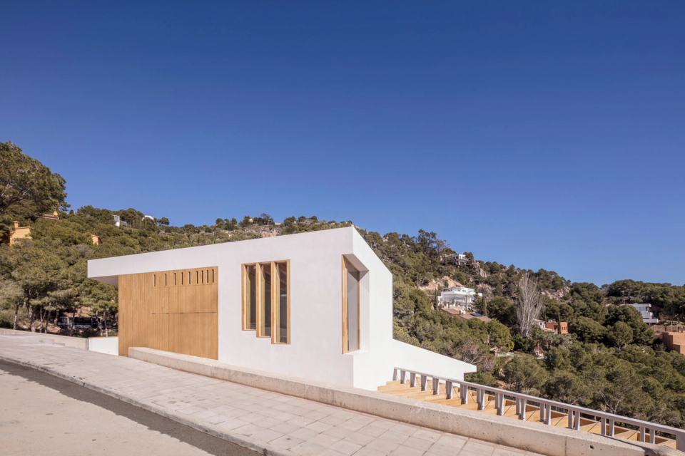 Detached single-family home naturally integrated into a steeply sloped plot surrounded by Mediterranean vegetation. The architecture combines white volumes, large glazed surfaces, and wood cladding to create a contemporary, warm, and landscape-oriented proposal. The project takes advantage of the site’s topography to generate different platforms and outdoor spaces visually connected to the natural surroundings and panoramic views. This detached house stands out for its balanced relationship between architecture, nature, and light, enhancing a peaceful residential experience integrated into the landscape.