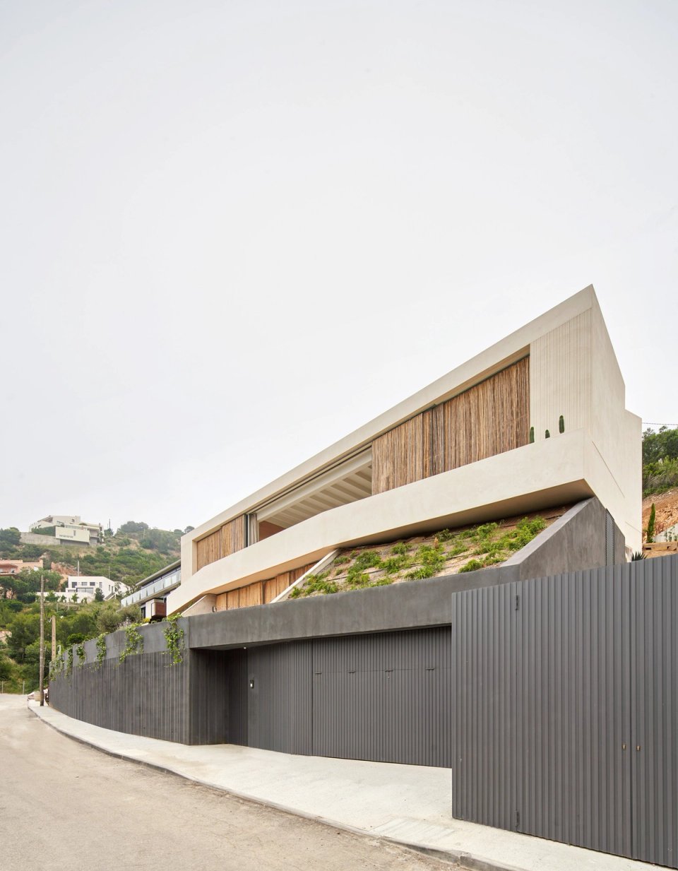 This single-family home stands out for its contemporary architecture that naturally adapts to the site's topography and the surrounding landscape. The horizontal composition of the volumes reinforces the feeling of integration with the mountain, while the large surfaces clad in natural materials provide warmth and texture to the whole. The dark base of the building acts as an element of containment and privacy, contrasting with the light-toned upper volumes that appear to float above the terrain. This single-family home combines minimalist design, integrated vegetation, and a fluid relationship between architecture and the Mediterranean environment.