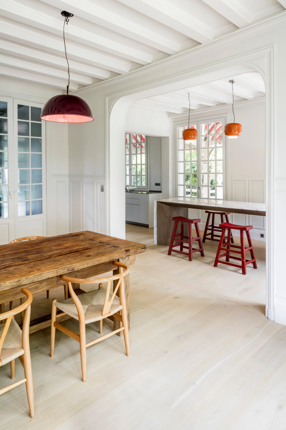 This interior renovation proposal stands out for the brightness and spaciousness of its spaces, combining a modern minimalist kitchen with premium materials such as wood and veined marble. Large windows with classic carpentry allow natural light to enter and create a visual connection with the exterior, while orange pendant lamps add a contemporary and sophisticated touch to the overall composition.