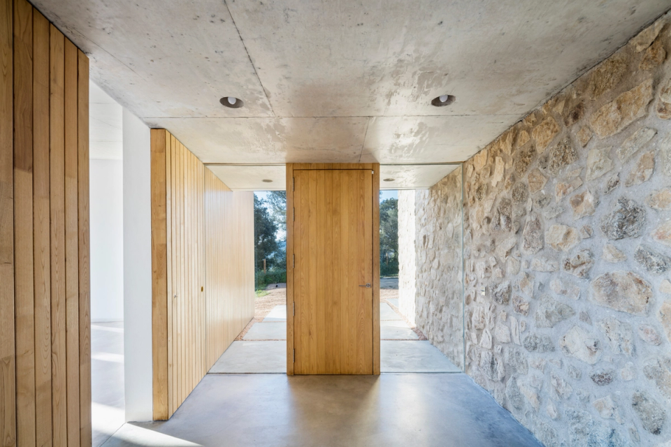 Interior connection space in a comprehensive home renovation project featuring architecture that combines natural materials with contemporary lines. Restored stone walls coexist with wooden claddings and exposed concrete ceilings, creating a warm, minimalist, and timeless atmosphere. Natural light flows through the space thanks to large openings and glass enclosures, enhancing the relationship between interior and exterior. This full renovation stands out for the quality of the finishes, the balance between tradition and modernity, and a layout designed to provide spaciousness, brightness, and comfort.