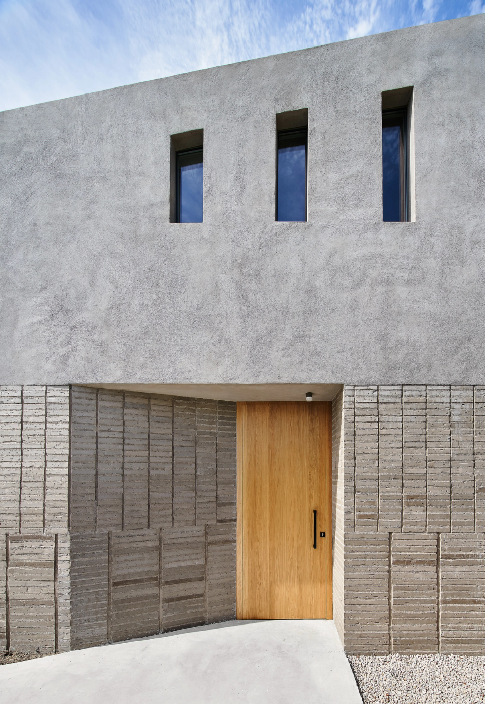 The entrance to this single-family home is resolved with a serene and timeless architecture, marked by the combination of mineral cladding, textured ceramics, and natural wood. Vertical openings and continuous lines reinforce the sense of order, light, and integration with the landscape.