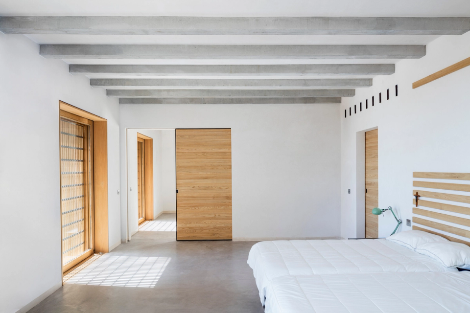 Master bedroom of a comprehensive renovation with a minimalist design that combines natural light, warm materials, and contemporary architecture. Exposed concrete beams, natural wood enclosures, and continuous flooring create a serene, elegant, and functional space. Large openings with solar protection filter the exterior light, generating a comfortable and intimate atmosphere. This full renovation embraces spacious and refined interiors where the simplicity of the finishes and the quality of the materials provide comfort, harmony, and a timeless aesthetic.