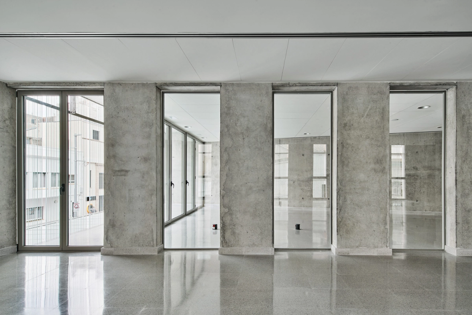 Interior space of a cultural center with minimalist and contemporary architecture defined by exposed concrete, large glazed surfaces, and natural light. The rhythmic composition of the vertical openings creates an orderly and elegant image, providing visual depth and connection with the exterior. This cultural center focuses on spacious, functional, and multipurpose spaces designed to encourage cultural, social, and community activities in a modern and bright environment.
