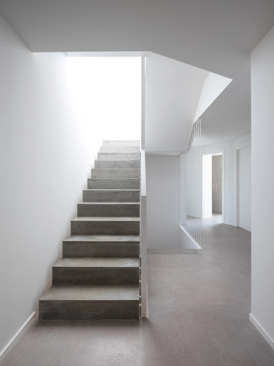 Interior detail of a contemporary detached single-family home where the staircase becomes a sculptural element integrated into the architecture of the space. Vertical metal railings, natural light, and a monochromatic palette reinforce a minimalist and elegant aesthetic, creating a sense of visual lightness and depth. The geometric composition and controlled entry of light enhance the sober and sophisticated character of the home, with an architecture designed to prioritize brightness, spatial continuity, and interior comfort.