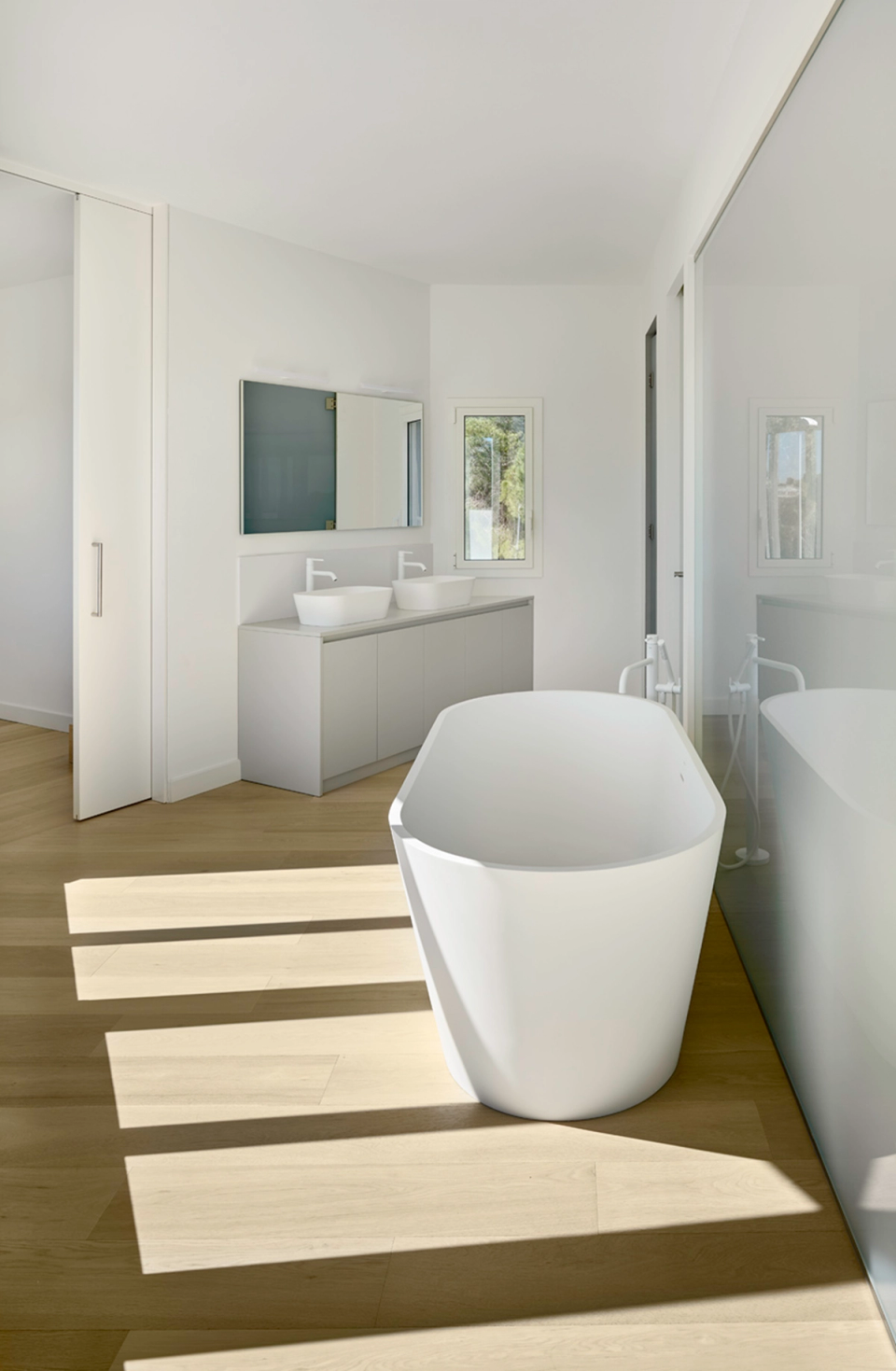 Main bathroom in a detached single-family home with minimalist design, freestanding bathtub, and contemporary finishes in light tones. The space stands out for its natural light, warm wooden flooring, and elegant layout that enhances comfort and functionality.