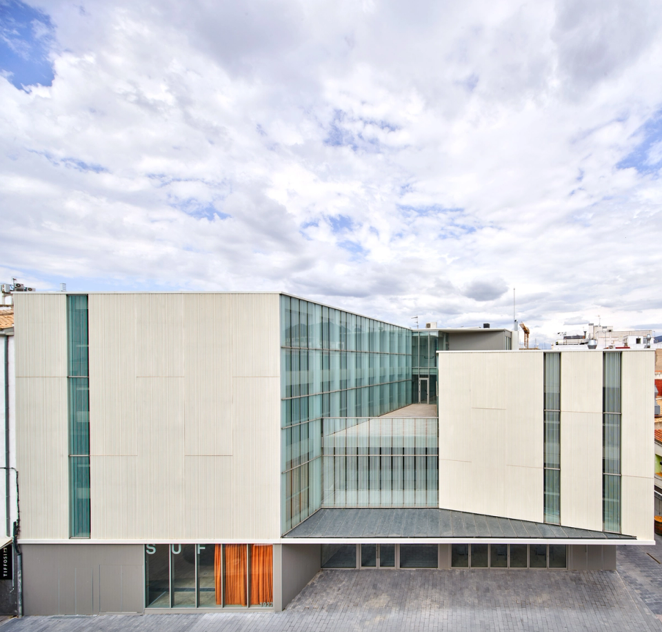 General view of a contemporary cultural center integrated into the urban fabric, with architecture defined by the combination of clean volumes, translucent façades, and large glazed surfaces. The building is organized around a central courtyard that encourages the entry of natural light and visual connection between spaces, creating an elegant, open, and functional image. Light finishes and minimalist geometry reinforce the institutional and cultural character of the facility.