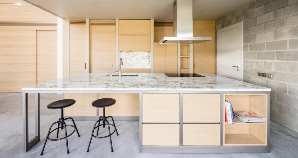 Open-plan minimalist kitchen conceived as the center of the living area. The white marble island combines with light wood furniture and metal structures, creating an elegant and contemporary contrast. Continuous concrete flooring and exposed walls add a soft industrial character, while the abundant natural light enhances the feeling of spaciousness, warmth, and connection with the rest of the home.