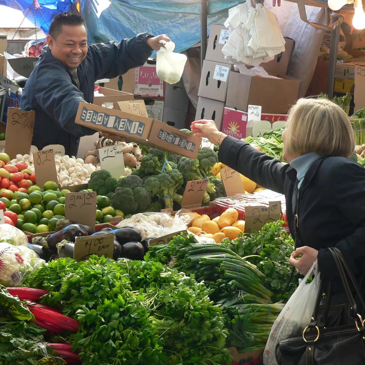 A Haymarket produce vendor smiling as he hands a purchase to a customer across a colorful display of vegetables