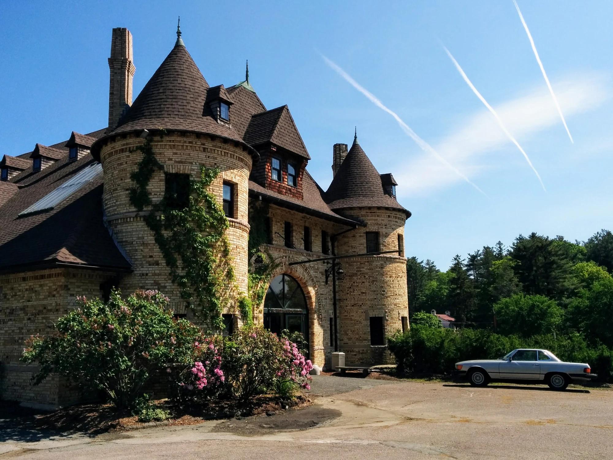 Turrets and an arched entrance at the Larz Anderson Auto Museum carriage house in Brookline, with flowering shrubs and a silver car outside.