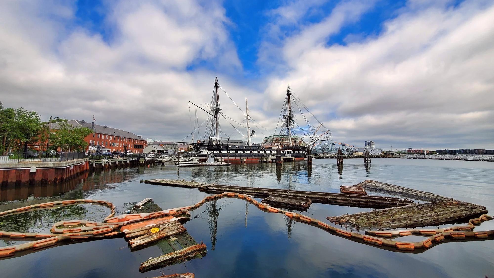 USS Constitution moored at Charlestown Navy Yard, with brick waterfront buildings and floating timber structures in the foreground.