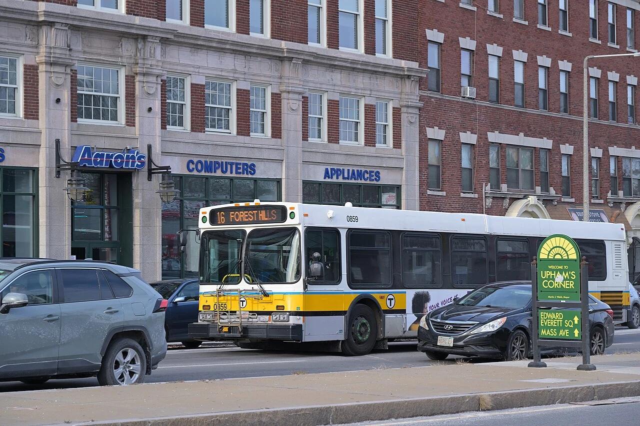 An MBTA bus passes brick storefronts and the Uphams Corner sign on Columbia Road in Dorchester.