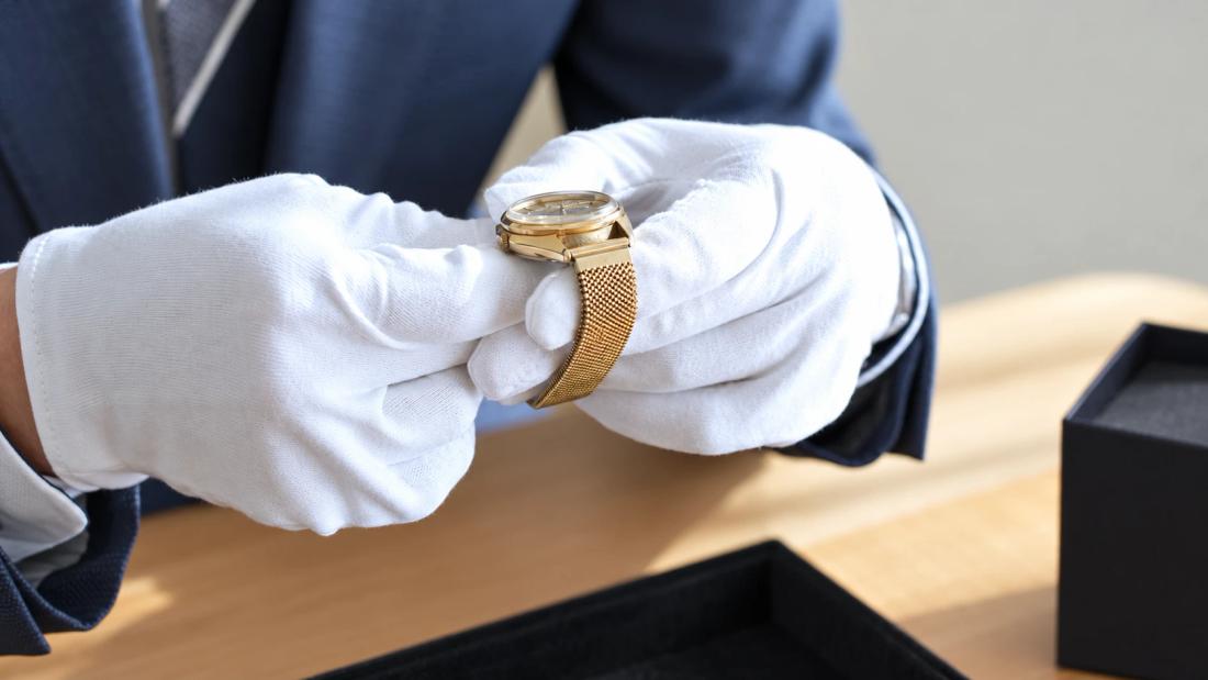 A specialist in white gloves holding a gold watch over a tray