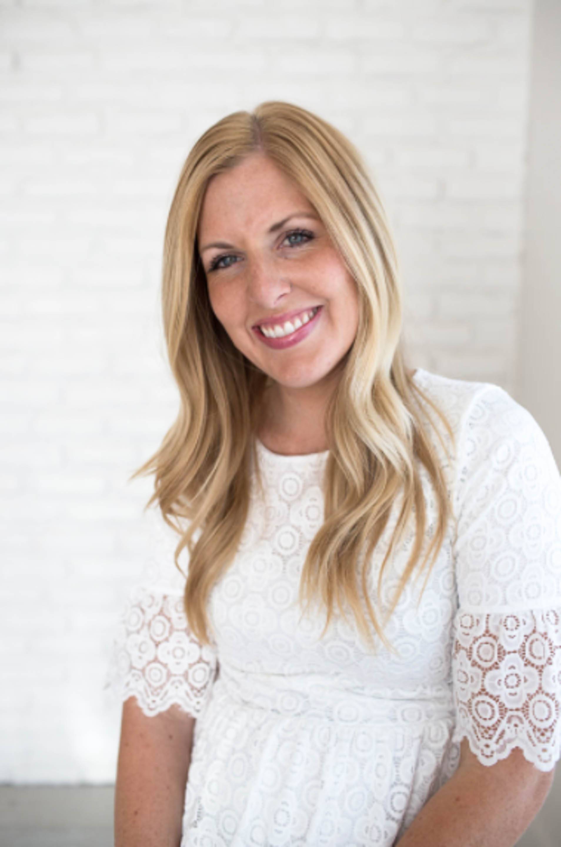 Smiling blonde woman wearing a white lace dress against a light brick wall background