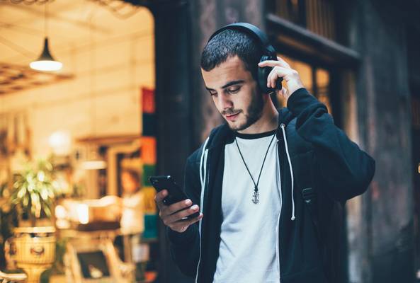 Young man wearing headphones and using smartphone outdoors near a cafe at night