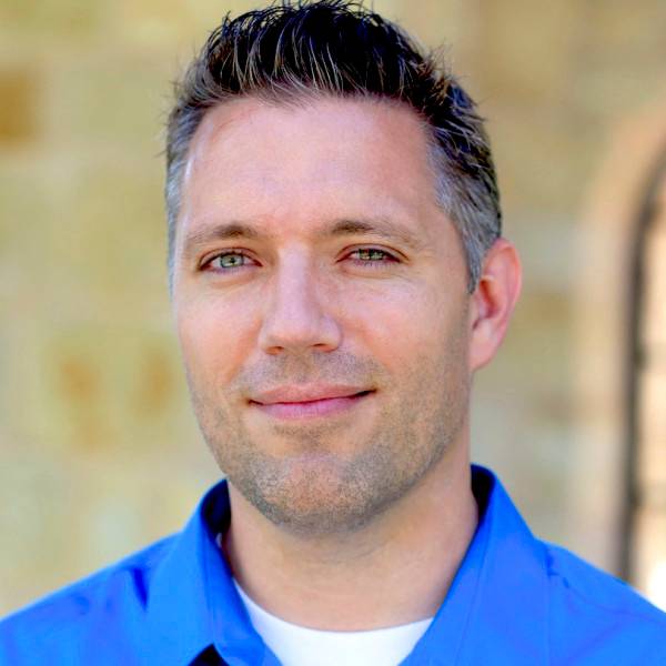 Close-up of a man with short hair wearing a blue shirt, smiling softly outdoors.