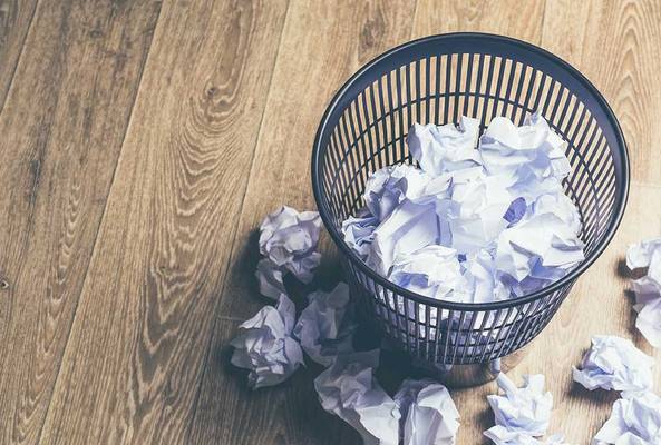 Black wastebasket filled with crumpled paper and more scattered on wooden floor.