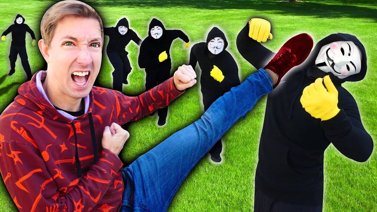 Man in red hoodie kicking multiple masked figures in black suits on grass field.