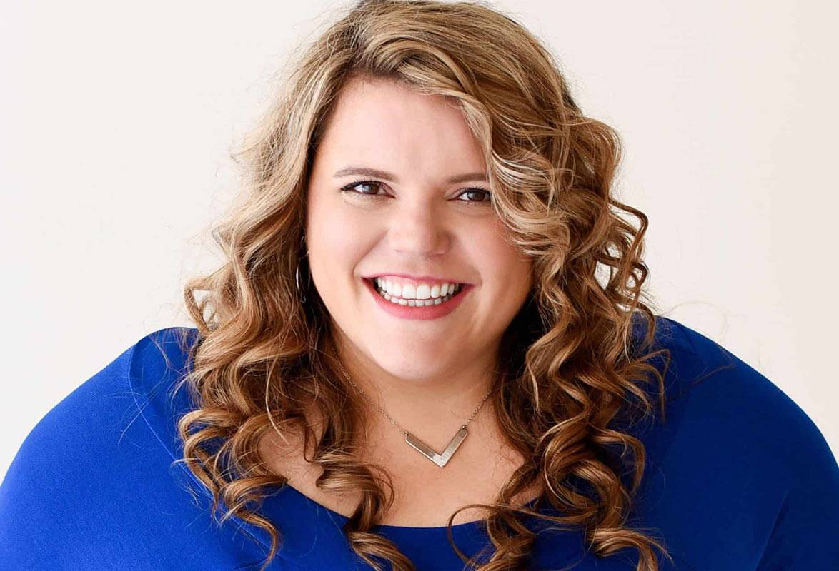 Smiling woman with curly hair wearing blue top and silver necklace on white background