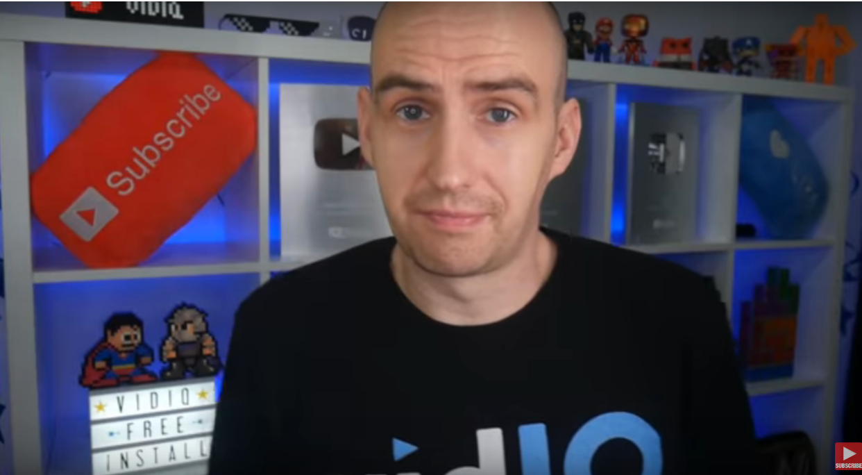 Man in black shirt in a video creator studio with subscribe pillow and YouTube awards.