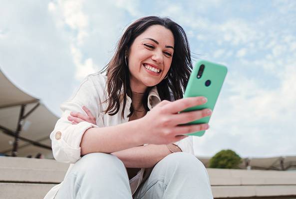 Smiling woman sitting outdoors using smartphone, enjoying social media or messaging.