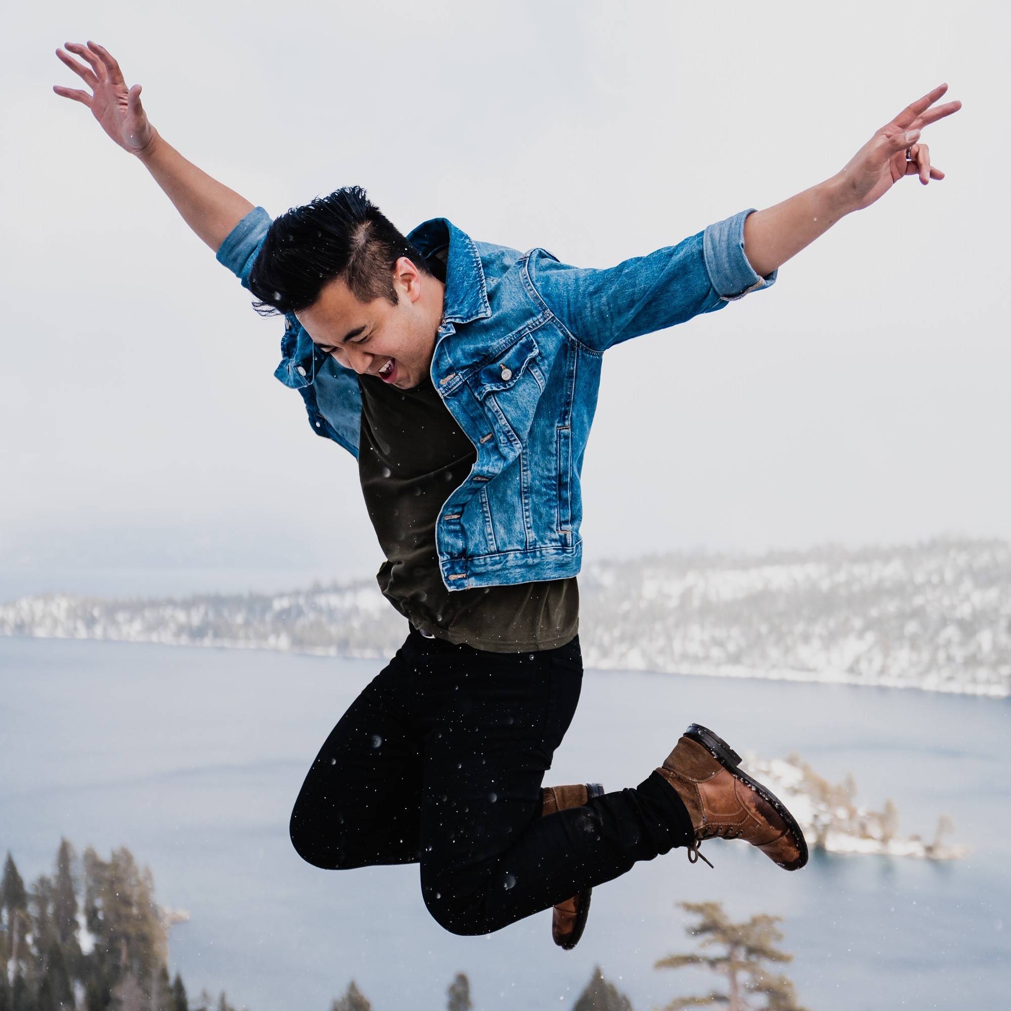 Young man in denim jacket joyfully jumping outdoors near snowy lake and trees