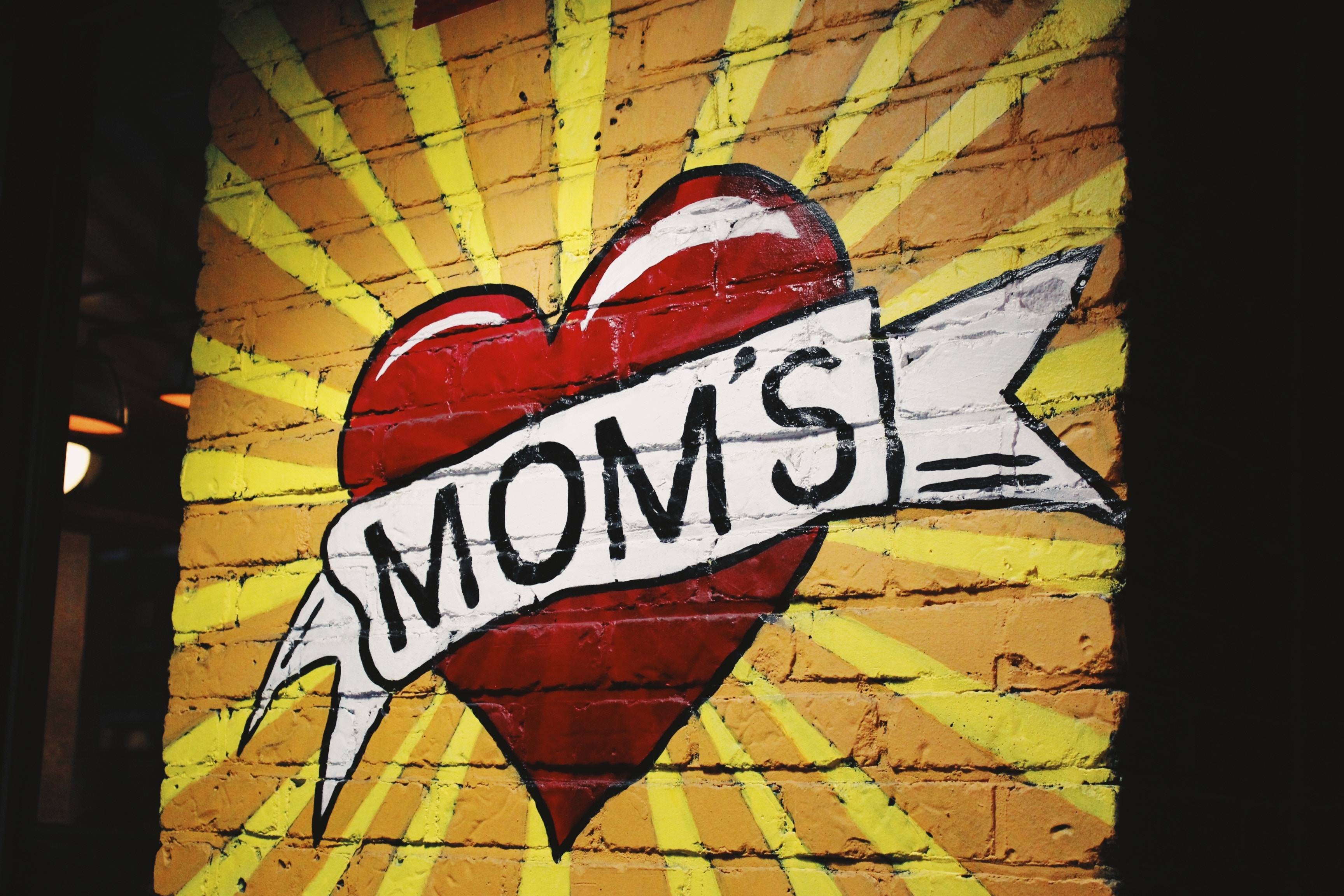 Red heart graffiti with "Mom's" banner on yellow brick wall background mural.