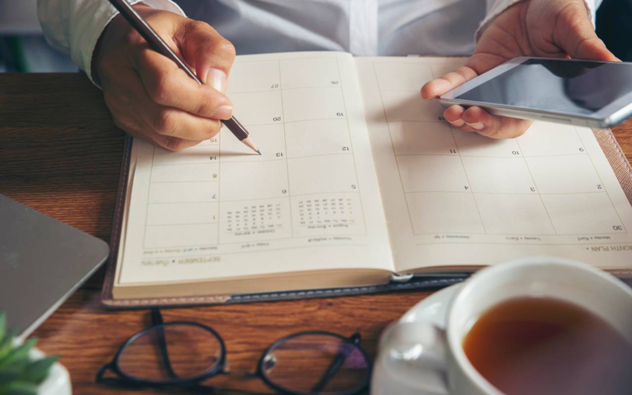 Person scheduling in planner with pencil and holding smartphone beside coffee cup