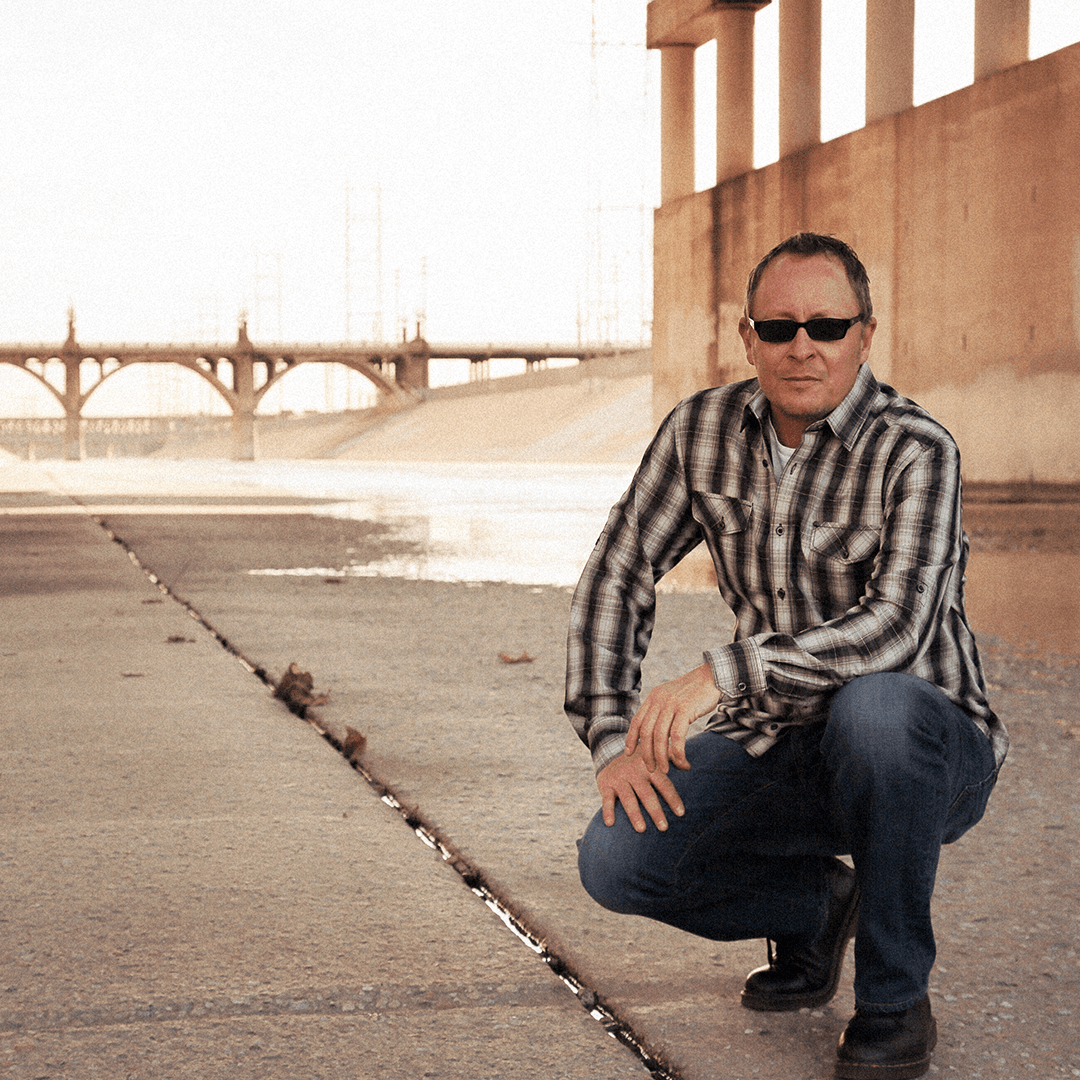 Man in sunglasses crouching on concrete under a bridge with an arched overpass in background