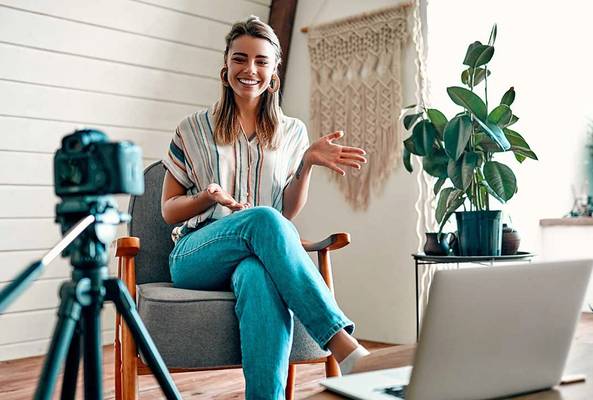 Young woman recording video blog sitting with camera and laptop indoors with plants.
