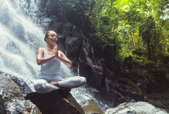 Woman meditating in yoga pose near waterfall surrounded by lush forest nature.