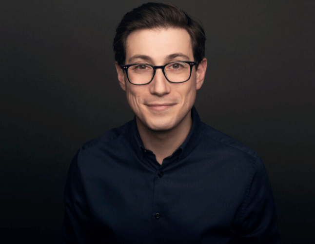 Young man wearing glasses and a dark shirt smiling against a black background