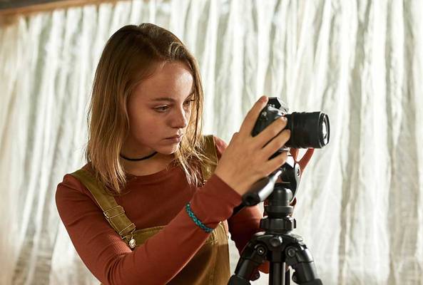Young woman adjusting a DSLR camera on a tripod indoors near sheer curtains